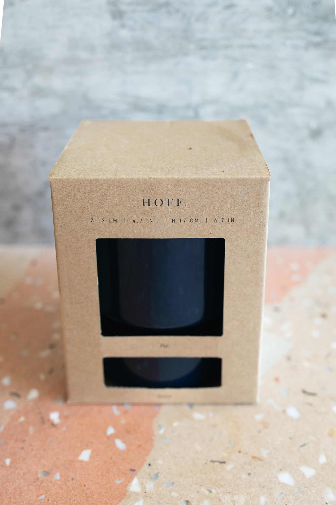 A kraft paper box with a clear window displaying a dark blue plant pot, labeled 'HOFF', set on a terrazzo surface against a textured gray background.