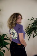 Person wearing a purple t-shirt with a graphic design of the Cancer Zodiac, standing indoors with plants in the background.