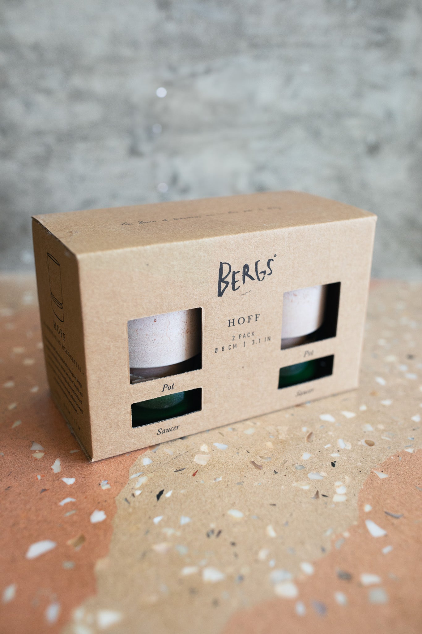 A kraft paper box of Berks Hoff 2 pack planter pots with saucers, showing through cutout windows, placed on a terrazzo surface against a textured gray background.