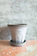 A small handmade concrete planter pot with a matching saucer, featuring a textured speckled finish and embossed arch details, set on a terrazzo surface against a textured gray wall.