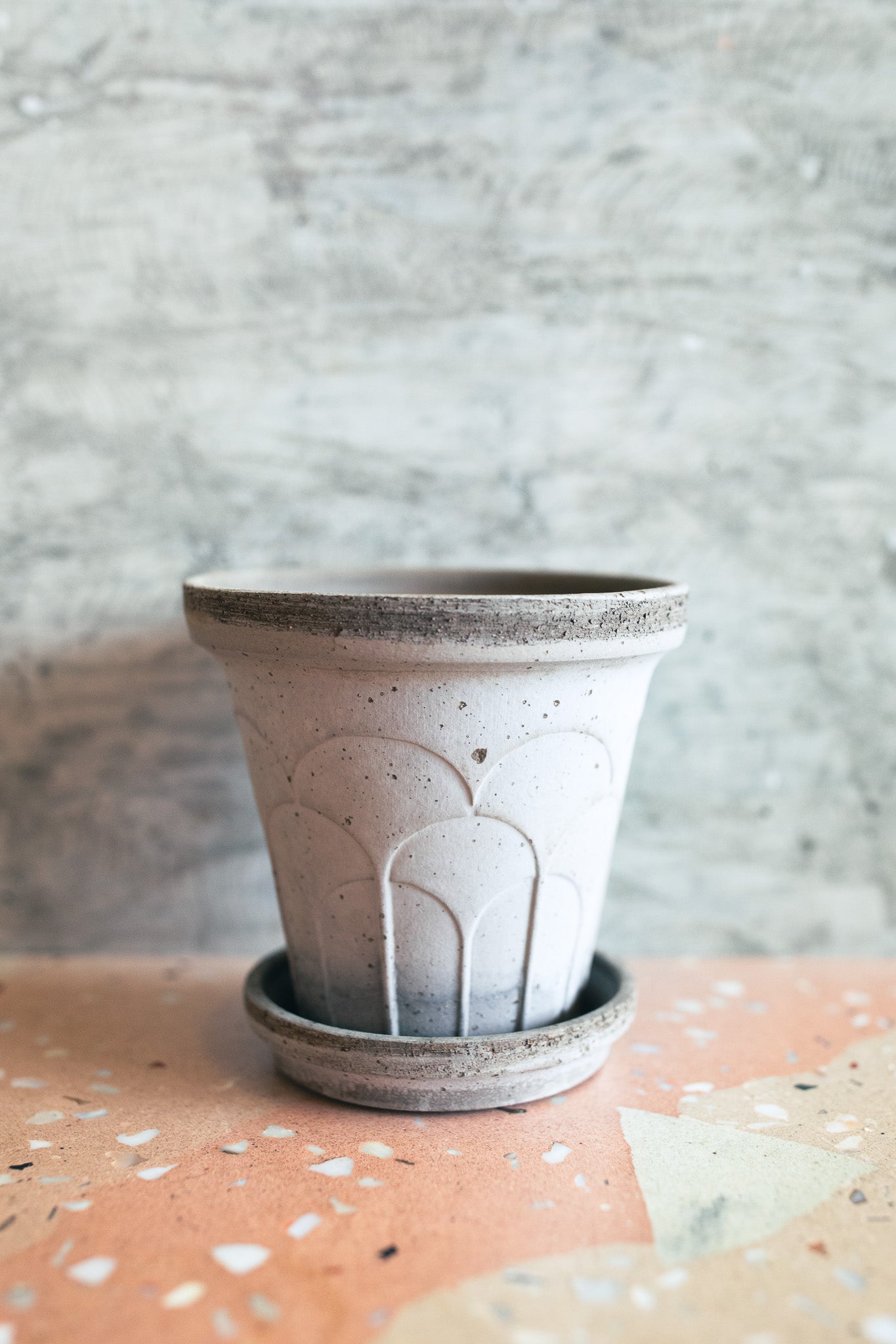 A small handmade concrete planter pot with a matching saucer, featuring a textured speckled finish and embossed arch details, set on a terrazzo surface against a textured gray wall.