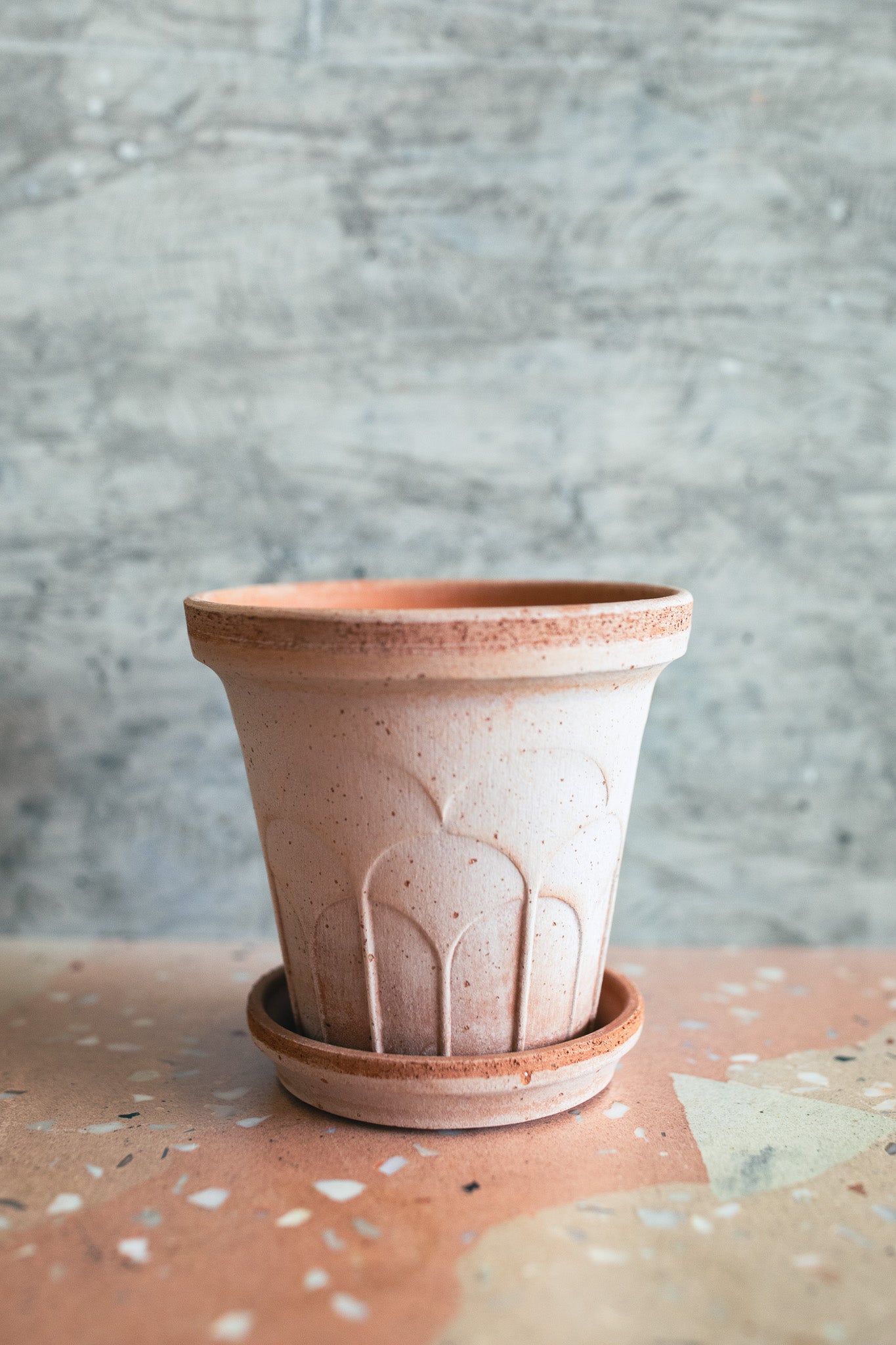 A small terracotta-colored ceramic planter pot with a matching saucer, featuring a textured embossed design on the front, set against a gray textured background.