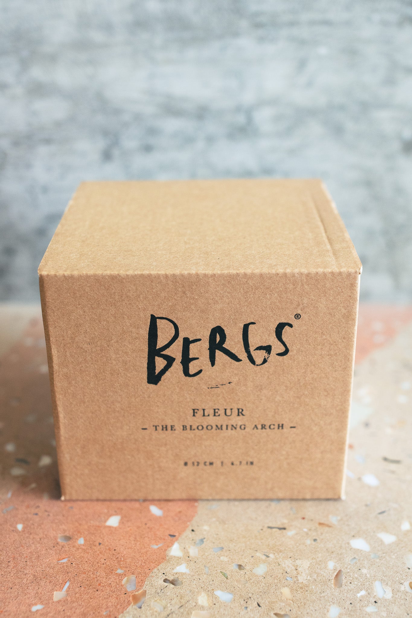 A square kraft paper box with 'BERGS' and 'FLEUR - THE BLOOMING ARCH -' printed on the front, photographed on a terrazzo-like surface against a textured gray wall.